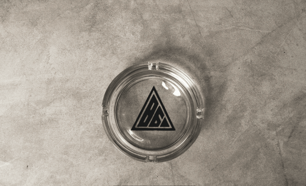 A transparent Glass Ashtray with a black triangular logo featuring stylized letters in its center, placed on a textured surface. This sleek stoner Glass Ashtray by Eighty Six Brand perfectly complements your collection of hemp derivatives.